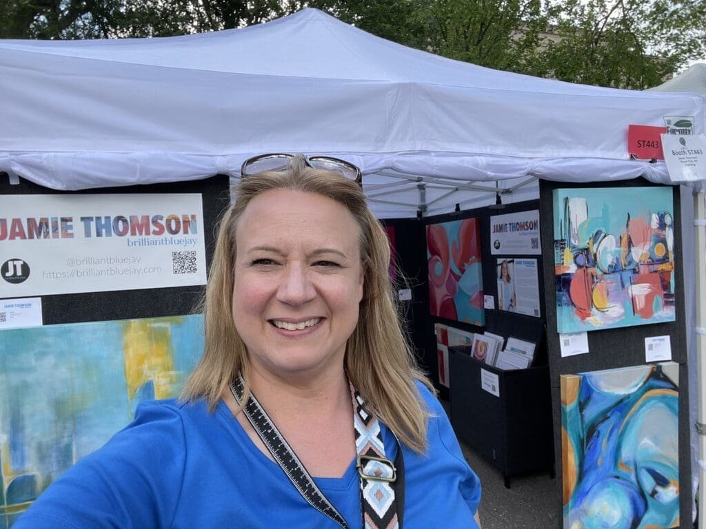 A woman in a blue shirt smiles in front of an art booth at the 2023 Funky Ferndale Art Fair, displaying colorful abstract paintings beneath a white canopy marked JAMIE THOMSON and brilliantbluejay. Trees frame the lively scene.