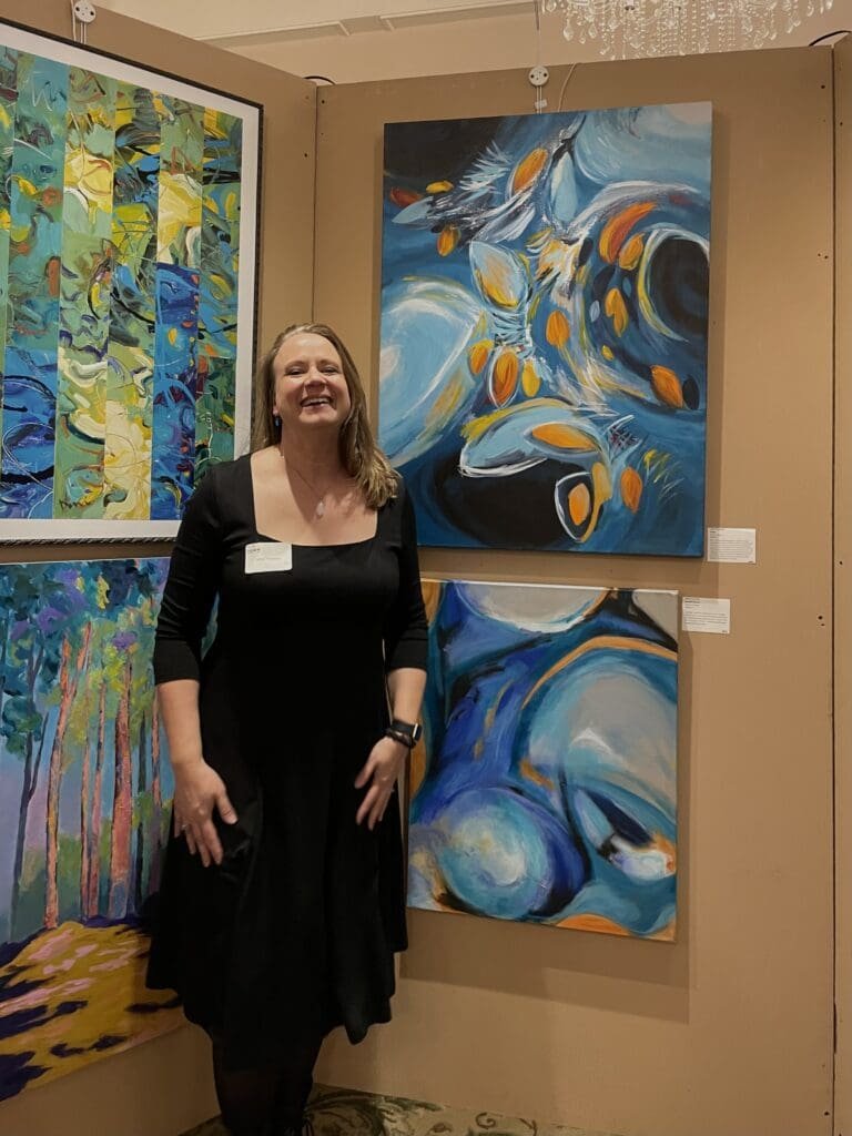 A woman in a black dress stands smiling in front of colorful abstract paintings displayed on a tan wall at the Our Town 2024 art exhibit.
