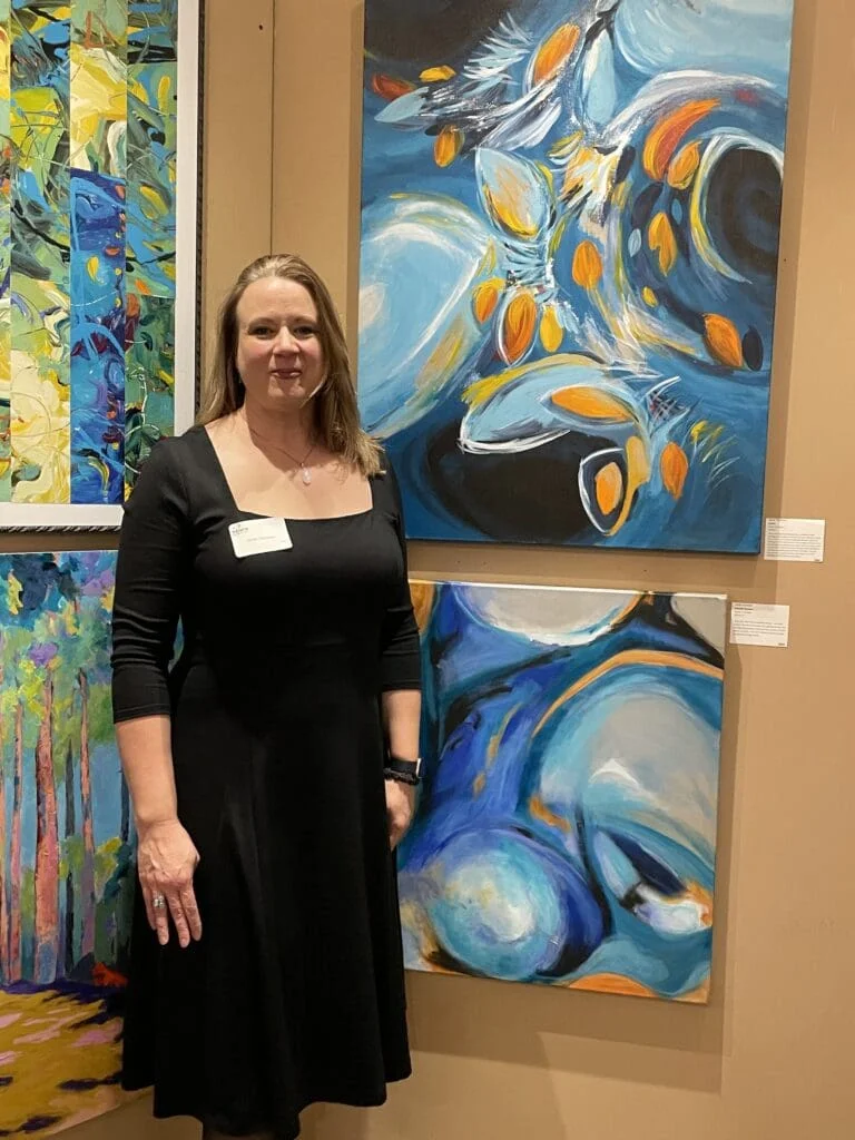 A woman in a black dress stands beside colorful abstract paintings displayed on the gallery wall at the 2024 Our Town exhibit. She faces the camera, wearing a white name tag.