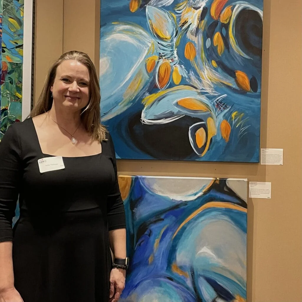 A woman in a black dress stands smiling beside abstract paintings with blue, orange, and white colors on display in an art gallery during the Our Town 2024 play.