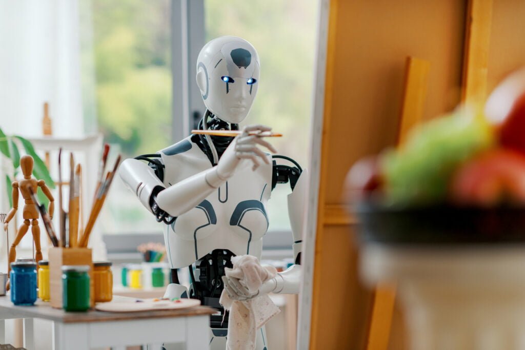 A humanoid robot with blue-lit eyes holds a paintbrush and cloth, painting at an easel in a bright studio. Art supplies and a bowl of fruit suggest a still life scene, blending art and AI in the creative process of the digital age.