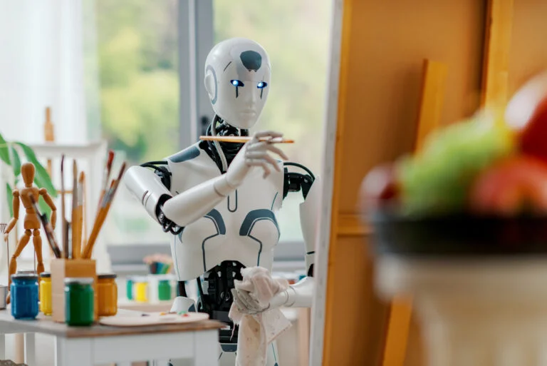 A humanoid robot with blue-lit eyes holds a paintbrush and cloth, painting at an easel in a bright studio. Art supplies and a bowl of fruit suggest a still life scene, blending art and AI in the creative process of the digital age.