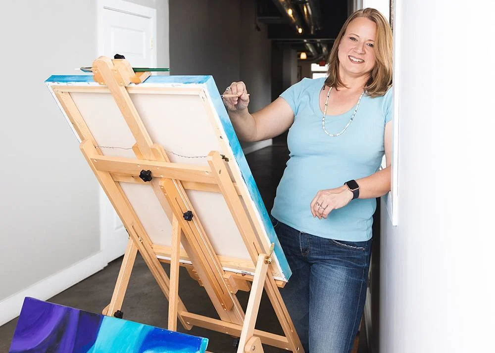 A smiling woman in a light blue shirt stands next to a white wall, painting on a canvas set on an easel in a hallway. She holds a brush and appears relaxed and happy.